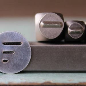 May include: A close-up shot of metal stamping tools. A round metal stamp with three horizontal lines is in the foreground. Three square metal stamps with rounded tops are in the background. The tools are on a dark gray surface.