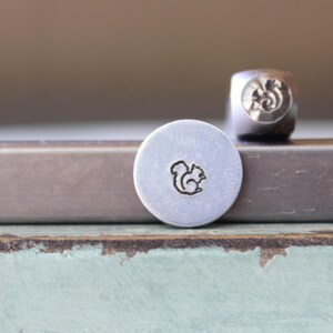 May include: A metal stamp and a stamped metal disc. The stamp is a small, square block with a squirrel design on one end. The disc is round and has a squirrel design stamped on it. The squirrel design is a simple outline.