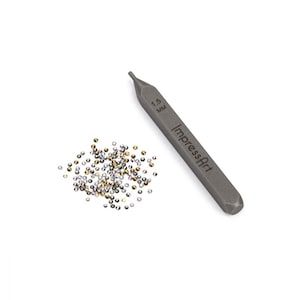 May include: A gray metal tool with the text "ImpressArt" and "1.5 MM" engraved on it. A pile of small, clear, silver, and gold rhinestones are scattered on a white background.