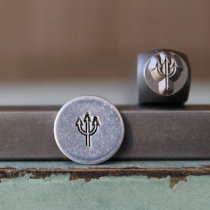 May include: A close-up of a metal stamp with a trident design. The stamp is silver and has a black trident design. The stamp is sitting on a gray surface.