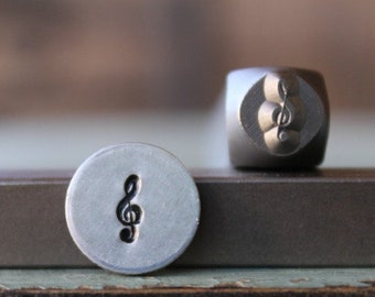 May include: Two metal stamps, one round and one cube-shaped, each featuring a treble clef symbol. The round stamp has a clear, stamped image of the clef. The cube stamp has the clef design on its face. The stamps are on a dark surface.