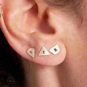Puede incluir: Tres pendientes de oro en forma de triángulo con las letras B, E y N grabadas. Los pendientes se llevan en el lóbulo superior de la oreja.