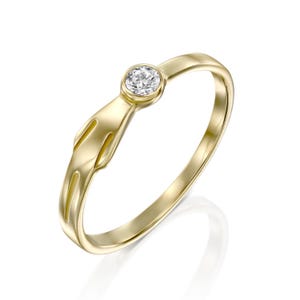 Op de afbeelding: Een gouden ring met een enkele, ronde, heldere edelsteen. De ring heeft een uniek ontwerp met een gestileerd, opengewerkt detail op de band. De ring staat tegen een witte achtergrond.