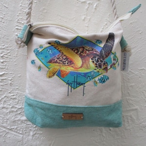 Puede incluir: Bolso bandolera de lona blanquecina con base turquesa y correa de cuerda. El bolso presenta un gráfico colorido de una tortuga marina dentro de un marco en forma de diamante, rodeado de peces. Cierre con cremallera.