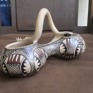 以下が含まれることがあります： 黒と白の幾何学模様に赤いアクセントが施された、2つのボウルが付いたセラミック製のサービングディッシュ。ハンドルは、スタイリッシュな目をした蛇の形をしています。