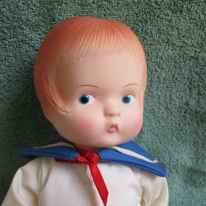 Puede incluir: Una muñeca de plástico vintage con cabello rojo, ojos azules y mejillas sonrosadas. La muñeca lleva una camisa de marinero blanca con cuello azul y una corbata de lazo roja.