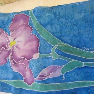 May include: A hand-painted silk scarf featuring a purple iris flower with green leaves on a blue background. The artist's signature, "T.M.", is visible in the bottom right corner.