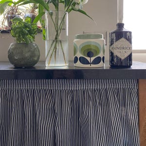 May include: A still life arrangement featuring a potted plant, a clear vase with flowers, a decorative jar with a floral design, and a dark gin bottle. A black and white striped curtain hangs below.