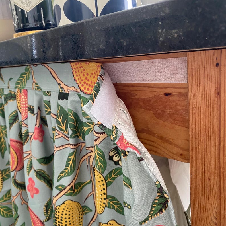 May include: A light blue fabric curtain with a repeating pattern of yellow lemons, red fruits, and green leaves. The curtain features a white hook-and-loop fastener strip along the top edge, which is attached to the wooden frame of a cabinet.