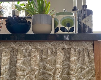 May include: A gathered fabric curtain featuring a repeating brown and white leaf pattern, positioned below a black countertop holding a dark blue bowl of grapes, a two-toned grey and beige planter with an aloe plant, a patterned canister, and a bottle.