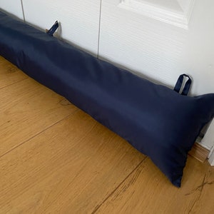 May include: A navy blue door draft stopper, a long cylindrical pillow, is placed at the bottom of a white door to prevent drafts. The door draft stopper is made of fabric and has two loops on each end.