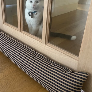 Può includere: Un fermaporta a strisce nere e beige con il testo "scandalo" e "london" sul lato. Un gatto bianco con macchie nere guarda attraverso una porta di vetro.