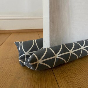 Könnte beinhalten: Ein grauer und weißer Türstopper mit geometrischem Muster. Der Türstopper ist aus Stoff und mit einem weichen Material gefüllt.