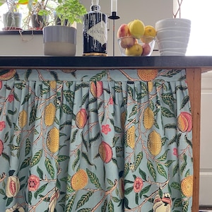 May include: A light blue fabric curtain with a repeating botanical print featuring yellow lemons, pink peaches, and green leaves. The curtain is gathered at the top with a straight header and hangs vertically to cover the base of a kitchen island.
