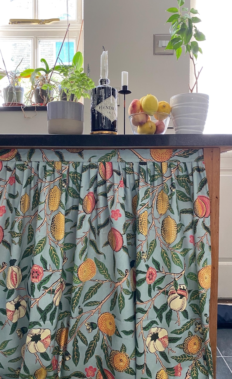 May include: A light blue fabric curtain with a repeating botanical print featuring yellow lemons, pink peaches, and green leaves. The curtain is gathered at the top with a straight header and hangs vertically to cover the base of a kitchen island.