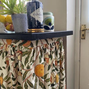 May include: A gathered fabric curtain with a cream background featuring a repeating botanical print of orange citrus fruits, green leaves, and brown branches. The curtain is attached to the underside of a black countertop surface.