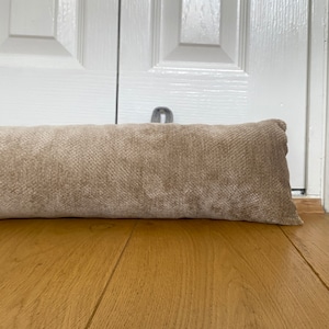 Può includere: Un fermaporta in tessuto beige con una piccola maniglia in metallo. Il fermaporta rettangolare è posto su un pavimento in legno davanti a una porta bianca. È progettato per impedire a una porta di aprirsi troppo o di sbattere.