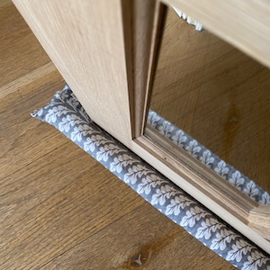 Könnte beinhalten: Ein grauer und weißer Türstopper mit einem Blattdesign. Der Türstopper befindet sich unter einer Holztür mit einem Glaseinsatz.