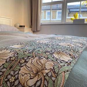 May include: A green, brown, and cream patterned bed runner with a floral design. The runner is on a light green bedspread.
