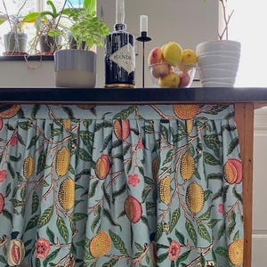 May include: A gathered fabric curtain featuring a light blue background with a repeating pattern of yellow lemons, pink peaches, green leaves, and small pink flowers. The curtain is attached to the front edge of a dark countertop surface.