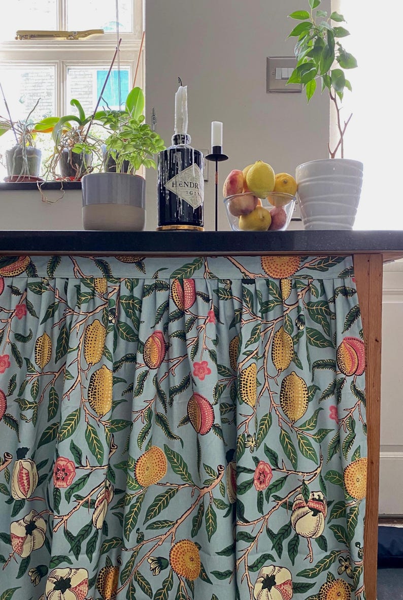 May include: A gathered fabric curtain featuring a light blue background with a repeating pattern of yellow lemons, pink peaches, green leaves, and small pink flowers. The curtain is attached to the front edge of a dark countertop surface.
