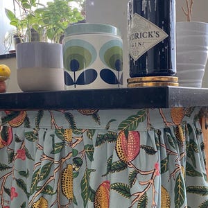 May include: A light blue fabric skirt with a repeating pattern of yellow and pink fruits and green leaves attached to a black surface. The fabric features gathered pleats along the top edge and covers the area beneath the black tabletop.