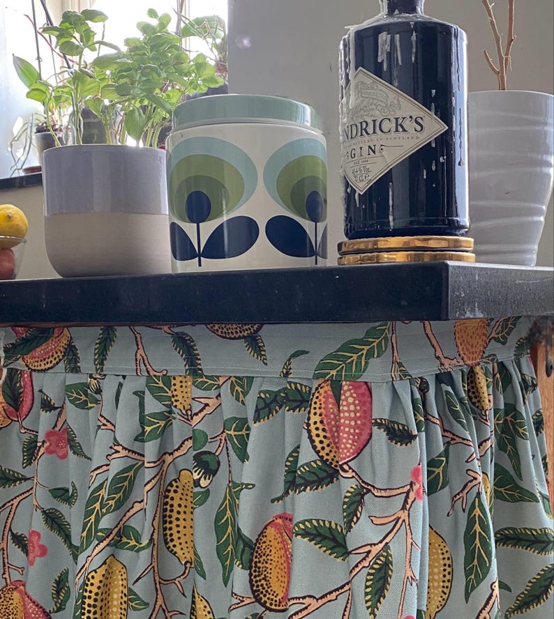 May include: A light blue fabric skirt with a repeating pattern of yellow and pink fruits and green leaves attached to a black surface. The fabric features gathered pleats along the top edge and covers the area beneath the black tabletop.