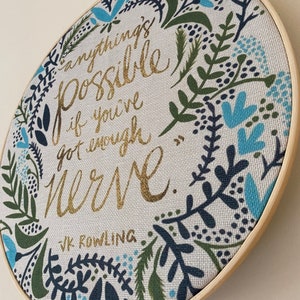 May include: A round embroidery hoop with a white fabric background and a gold quote that reads "Anything's possible if you've got enough nerve." The quote is attributed to J.K. Rowling. The fabric is decorated with a blue and green floral pattern.