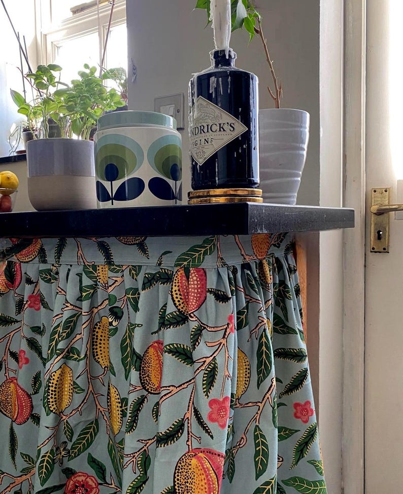 May include: A light blue fabric skirt with a repeating pattern of yellow and orange fruits, green leaves, and small pink flowers attached to a black shelf. On the shelf sits a dark blue Hendrick's Gin bottle with a white candle, a patterned canister, and plants.