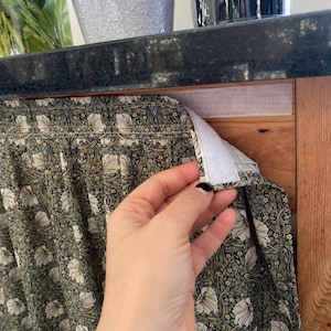Puede incluir: Una cortina de tela fruncida con un patrón botánico repetitivo de flores blancas y follaje verde sobre un fondo oscuro. La cortina tiene una tira de cierre de velcro blanca en el borde superior para fijarla a un marco de madera.