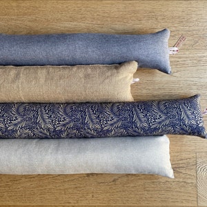 May include: Four doorstop pillows stacked on top of each other. The top doorstop is a dark blue denim fabric. The second doorstop is a light brown burlap fabric. The third doorstop is a dark blue fabric with a white floral pattern. The bottom doorstop is a light gray linen fabric.