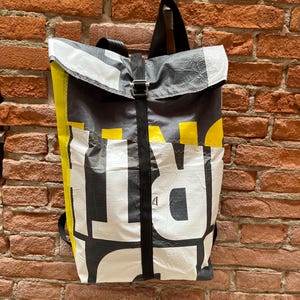 Può includere: Uno zaino con chiusura a rullo realizzato in materiale resistente alle intemperie, caratterizzato da una grafica audace in nero, bianco e giallo. Presenta una cinghia verticale nera centrale con chiusura a fibbia metallica e spallacci neri.