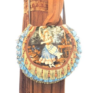 Pode incluir: Uma bolsa redonda de tape&ccedil;aria com franjas azuis e douradas. A tape&ccedil;aria apresenta uma mulher com um vestido azul e branco.