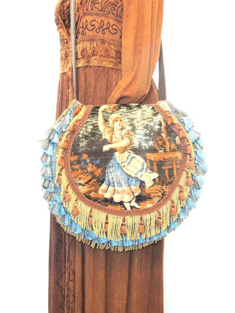 Pode incluir: Uma bolsa redonda de tape&ccedil;aria com franjas azuis e douradas. A tape&ccedil;aria apresenta uma mulher com um vestido azul e branco.