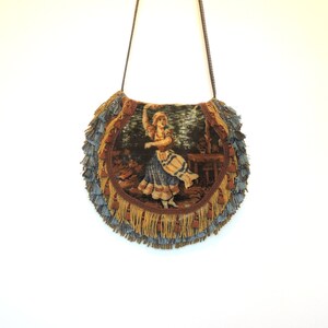 Pode incluir: Uma bolsa redonda de tape&ccedil;aria com uma borda de franjas em tons de marrom, dourado e azul. A tape&ccedil;aria apresenta uma ilustra&ccedil;&atilde;o vintage de uma mulher com um vestido azul dan&ccedil;ando.
