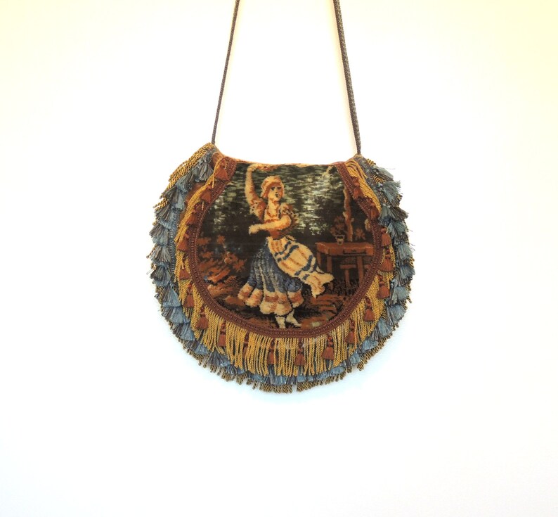 Pode incluir: Uma bolsa redonda de tape&ccedil;aria com uma borda de franjas em tons de marrom, dourado e azul. A tape&ccedil;aria apresenta uma ilustra&ccedil;&atilde;o vintage de uma mulher com um vestido azul dan&ccedil;ando.