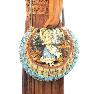 Pode incluir: Uma bolsa redonda de tape&ccedil;aria com franjas azuis e douradas. A tape&ccedil;aria apresenta uma mulher com um vestido azul em p&eacute; num jardim.