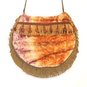 Pode incluir: Uma bolsa redonda de veludo laranja e rosa com acabamento em franjas douradas. A bolsa tem uma al&ccedil;a longa e &eacute; perfeita para carregar os itens essenciais do dia a dia.