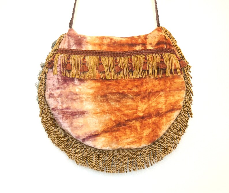 Pode incluir: Uma bolsa redonda de veludo laranja e rosa com acabamento em franjas douradas. A bolsa tem uma al&ccedil;a longa e &eacute; perfeita para carregar os itens essenciais do dia a dia.
