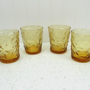 May include: A set of four amber-colored glass tumblers featuring a textured, wavy surface pattern. Each glass has a rounded rim and a slightly tapered base, showcasing a translucent, warm yellow hue with a distinct embossed ripple design.