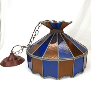 Op de afbeelding: Een hanglamp van gekleurd glas met een geometrisch patroon in blauw en bruin. De lamp heeft een ketting en een metalen kap.