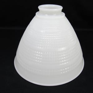 May include: A white milk glass lamp shade with a bell-shaped profile and a textured grid pattern consisting of small raised squares. The shade features a circular opening at the top with a raised collar for mounting, and a wide, smooth bottom rim.