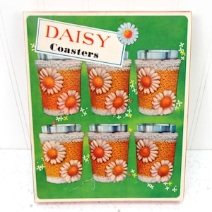 May include: A set of six daisy coasters in a box. Each coaster features a daisy design with orange and white petals on a textured orange background. The box has the words "DAISY Coasters" on a white and blue label, set against a green background.