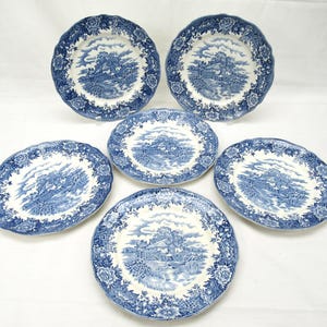 May include: A set of five round ceramic plates featuring a blue and white transferware pattern. Each plate displays a central landscape scene of a cottage near a stream, surrounded by a decorative border of floral motifs and scalloped edges.