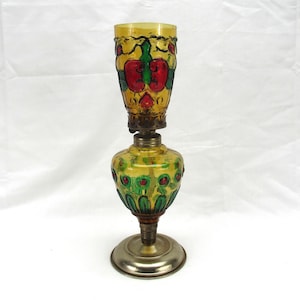 May include: An amber glass oil lamp featuring a hand-painted floral design in red and green. The lamp has a cylindrical top shade and a bulbous base, both decorated with painted motifs, connected by a metallic burner and a circular silver-toned base.
