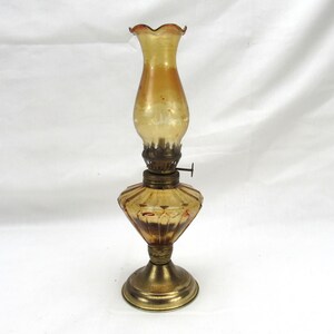May include: An amber-colored glass oil lamp featuring a fluted, ruffled chimney top and a faceted, diamond-patterned base. The lamp includes a metallic burner assembly with a small adjustment knob and a circular brass-toned pedestal stand.