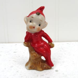 Peut inclure: Une figurine en céramique représentant un petit elfe avec un chapeau rouge pointu et une tenue rouge, assis sur une souche d'arbre marron. L'elfe a un visage pâle, de grandes oreilles et des traits peints, dont des yeux sombres et une petite bouche rouge, avec une finition brillante.