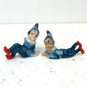 May include: Two ceramic figurines of elves in blue outfits with red shoes and hats. One elf is sitting, the other is lying down. Both have white collars and painted facial features.