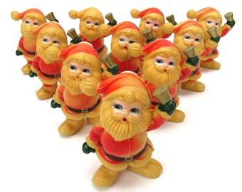 Può includere: Una collezione di dieci piccole statuette di Babbo Natale disposte in formazione triangolare. Ogni figura indossa un cappello e una giacca rossa con bordi gialli, una cintura nera e stivali gialli, tenendo in mano una campanella.