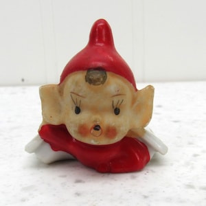 May include: A small ceramic figurine of a pixie-like character wearing a bright red pointed hat and a matching red collar. The figure has large ears, painted facial features including dark eyes and eyelashes, and a small round mouth.